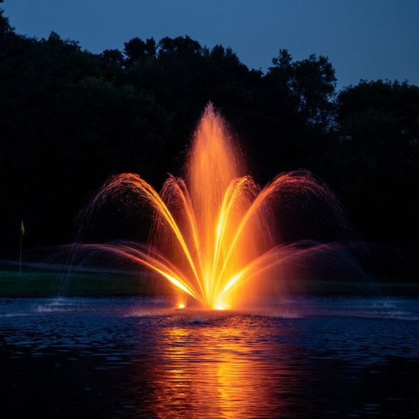 Fountain with orange water spray at night, reflecting on water surface with dark trees in background